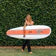 A orange and white Mega Magic model epoxy surfboard from Walden Surfboards carried by a woman walking on grass.