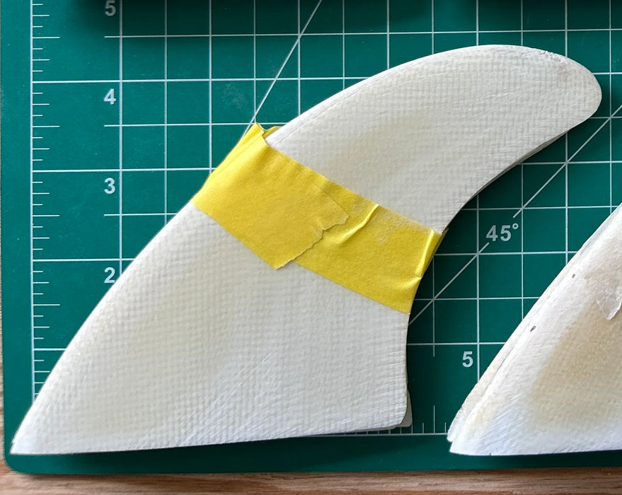 Three stacked on top of each other white surfboard fins with yellow tape on a green cutting mat with grid lines.