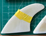 Three stacked on top of each other white surfboard fins with yellow tape on a green cutting mat with grid lines.