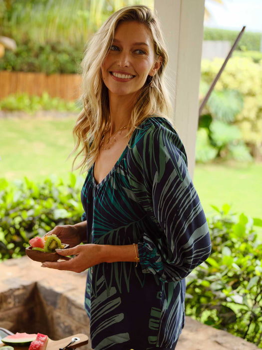 Woman wearing Phoebe Top in the Loulu Fronds print button front blouse with a scoop neckline holding food on a covered lanai in Hawaii.