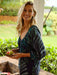 Woman wearing Phoebe Top in the Loulu Fronds print button front blouse with a scoop neckline holding food on a covered lanai in Hawaii.