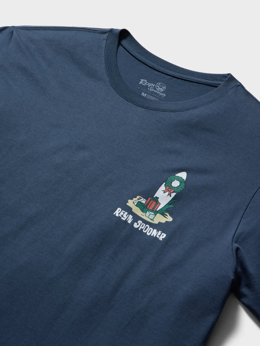 A close-up of a navy blue t-shirt front view featuring a small graphic on the left chest of a surfboard decorated with a green Christmas wreath and presents, with the text 'REYN SPOONER' printed below it.