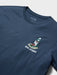 A close-up of a navy blue t-shirt front view featuring a small graphic on the left chest of a surfboard decorated with a green Christmas wreath and presents, with the text 'REYN SPOONER' printed below it.