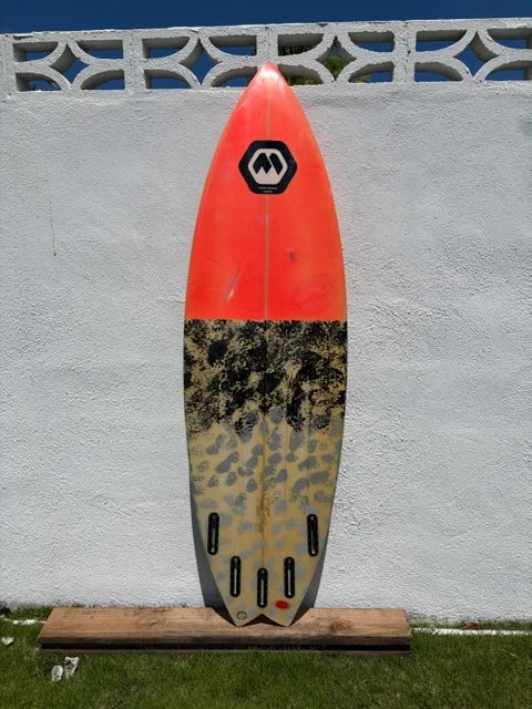 A shortboard surfboard bottom view with a bright orange and a grey/black camouflage patterned with 5 Futures fin boxes, displayed against a 6' white stucco wall.