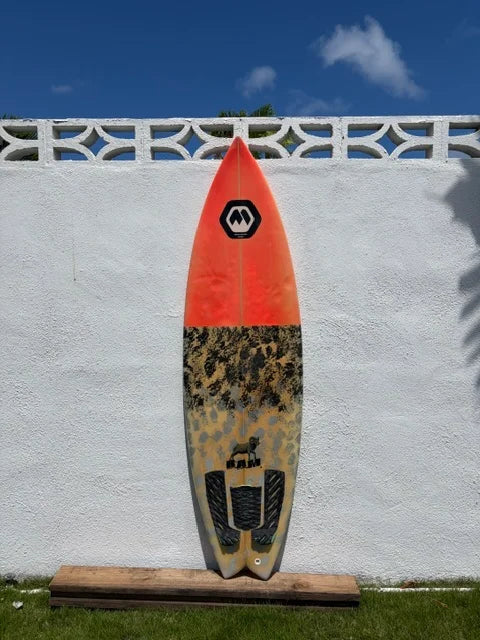 A shortboard surfboard deck view with a bright orange front and a grey/black camouflage patterned back, displayed  against a 6' white stucco wall outdoors.