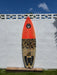 A shortboard surfboard deck view with a bright orange front and a grey/black camouflage patterned back, displayed  against a 6' white stucco wall outdoors.