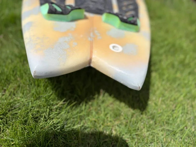 A close-up shot of a short board surfboard resting on green grass, showing its Swallow tail, a mottled yellow, gray, and white design, and green and black footpad.