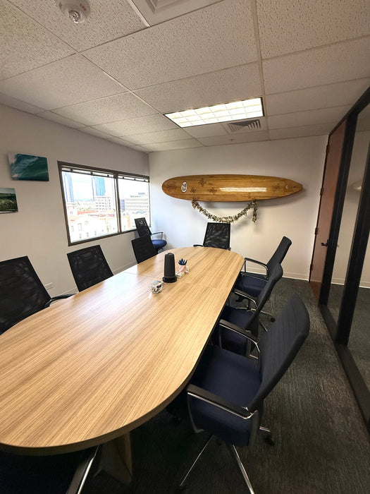 A modern office meeting room featuring a large, curved light-wood conference table surrounded by black mesh office chairs. A wooden surfboard is mounted on the white wall above and beyond the table as a decorative accent.