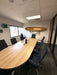 A modern office meeting room featuring a large, curved light-wood conference table surrounded by black mesh office chairs. A wooden surfboard is mounted on the white wall above and beyond the table as a decorative accent.