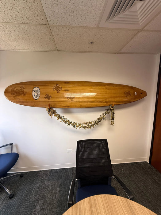 A wooden surfboard mounted on a white wall in an office setting, with a lei draped below it. A black mesh office chair is positioned in front of the board, with a blue office chair and a wooden table partially visible in the room.