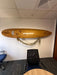 A wooden surfboard mounted on a white wall in an office setting, with a lei draped below it. A black mesh office chair is positioned in front of the board, with a blue office chair and a wooden table partially visible in the room.