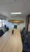 A modern office meeting room featuring a large, curved light-wood conference table surrounded by black mesh office chairs. With a zoom in on the wooden surfboard is mounted on the white wall above and beyond the table.