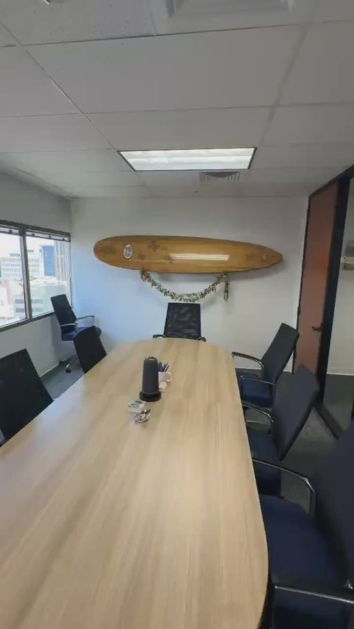A modern office meeting room featuring a large, curved light-wood conference table surrounded by black mesh office chairs. With a zoom in on the wooden surfboard is mounted on the white wall above and beyond the table.