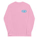 Pink long-sleeve crewneck shirt front view, with baby a blue and white graphic print of surfboards, palms and hut with the text 'KIMO'S SURF HUT' inside a double oval on the left chest.